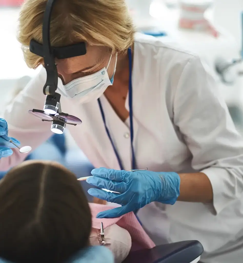 Das Bild zeigt eine Zahnärztin, die mit einer Lupenbrille und einem Zahnarztspiegel und weiteren Instrument in den Mund einer Patientin guckt. Die Zahnärztin trägt auch einen Mundschutz und Handschuhe. Das Bild wird im Bereich "Zahnreinigung (PZR)" verwendet.