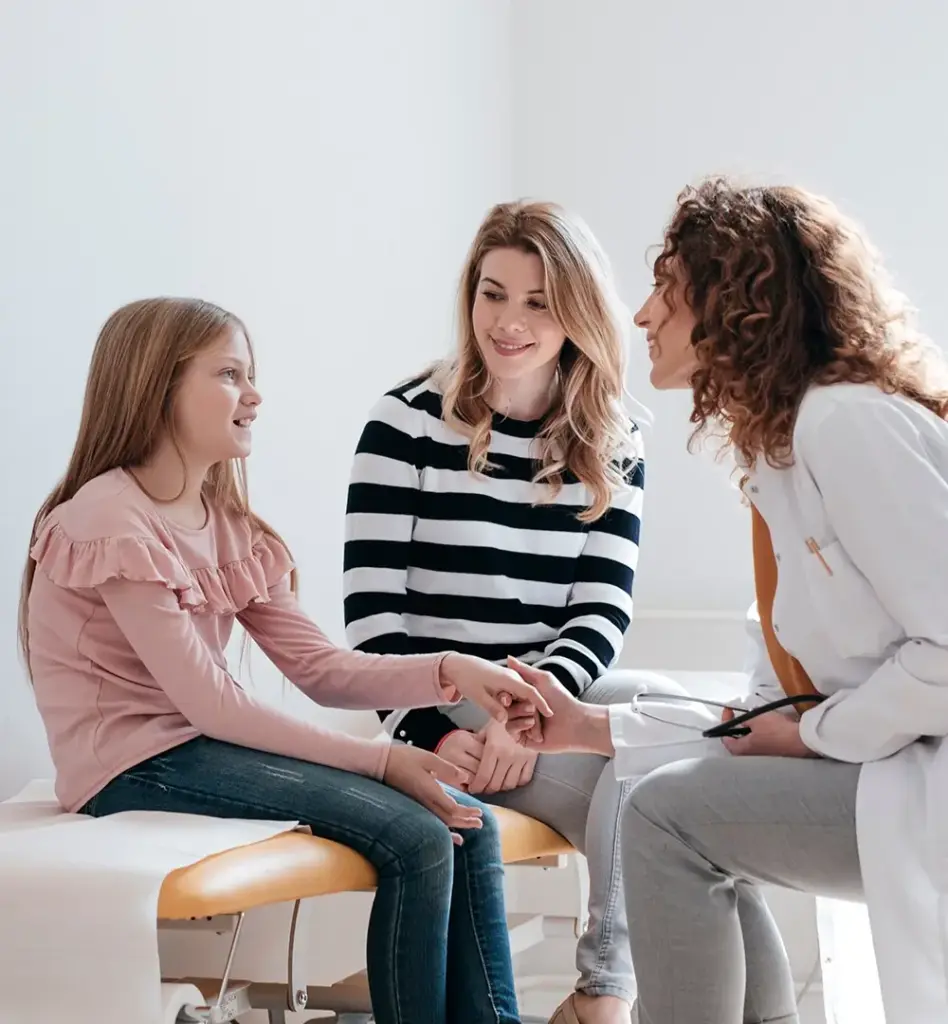Das Bild zeigt ein Mädchen, das auf einer Untersuchungsliege sitzt. Eine junge Frau sitzt neben ihr. Eine Ärztin hält eine Hand des Mädchens und lächelt es an. Das Bild wird im Bereich "Zusätzliche Vorsorgeangebote mit starke Kids by BKK" verwendet.