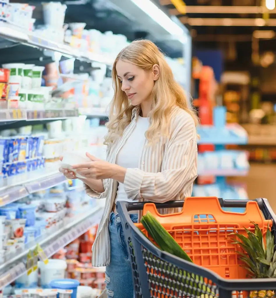 Eine Frau steht mit Einkaufswagen im Supermarkt und liest die Infos auf einem Produkt.
