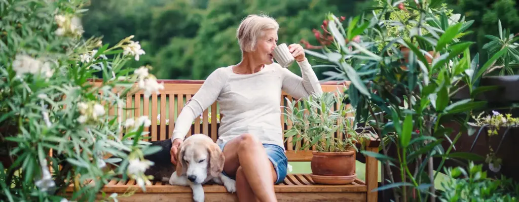 Eine Frau sitzt im Garten auf einer Bank und trinkt Kaffee. Neben ihr liegt ihr Hund.
