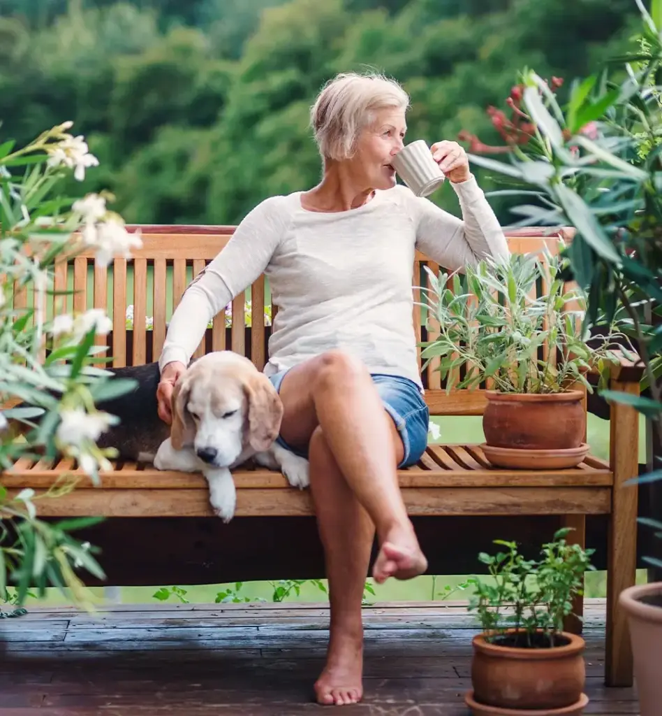 Eine Frau sitzt im Garten auf einer Bank und trinkt Kaffee. Neben ihr liegt ihr Hund.