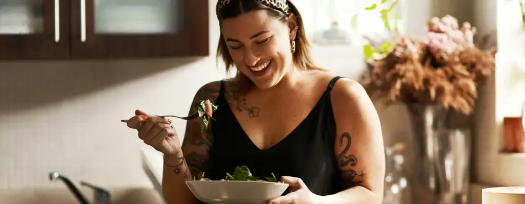 Eine Frau steht sommerlich gekleidet in einer Küche. Sie hält eine Schüssel und eine Gabel in den Händen und lächelt.