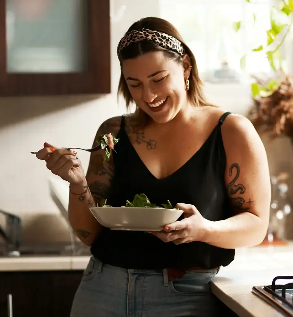 Eine Frau steht sommerlich gekleidet in einer Küche. Sie hält eine Schüssel und eine Gabel in den Händen und lächelt.