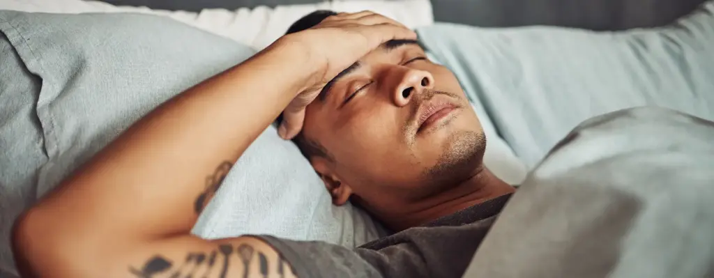 Ein Mann liegt im Bett, hat die Augen noch geschlossen und hält sich die Hand an die Stirn.