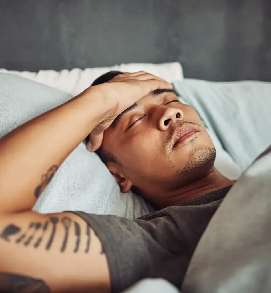 Ein Mann liegt im Bett, hat die Augen noch geschlossen und hält sich die Hand an die Stirn.
