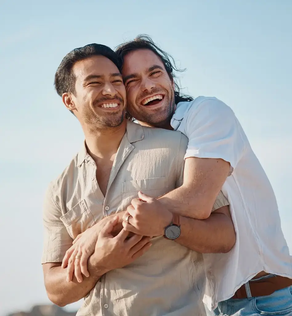 Zwei Männer stehen zusammen. Der eine Mann umarmt den anderen Mann von hinten. Sie beide lachen.