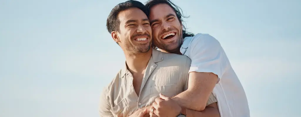 Zwei Männer stehen zusammen. Der eine Mann umarmt den anderen Mann von hinten. Sie beide lachen.