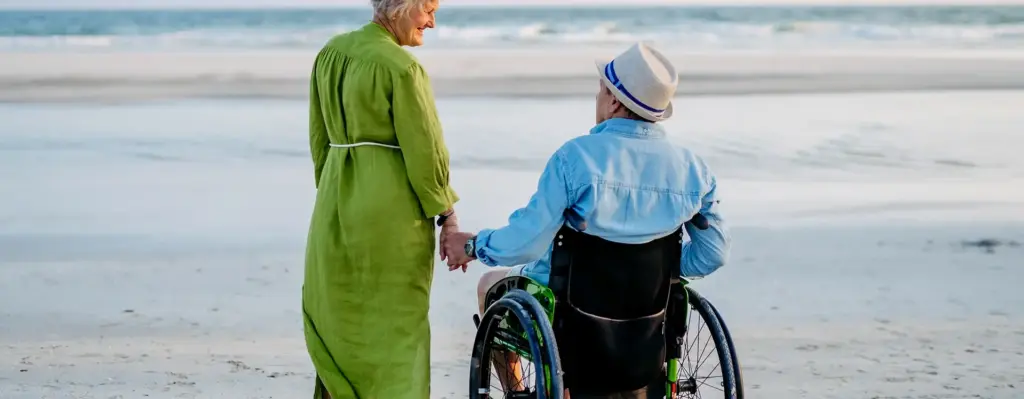 Das Bild zeigt einen älteren Mann mit Hut im Rollstuhl am Strand. Neben ihm steht eine Frau im grünen Kleid und hält seine Hand. Beide sieht man von hinten. Das Bild wird im Bereich "Aufenthalt im Ausland" verwendet.