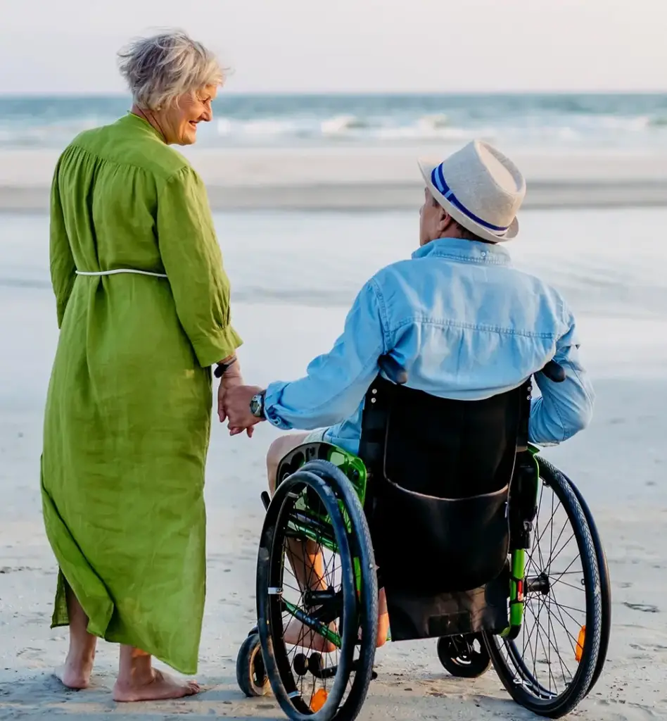 Das Bild zeigt einen älteren Mann mit Hut im Rollstuhl am Strand. Neben ihm steht eine Frau im grünen Kleid und hält seine Hand. Beide sieht man von hinten. Das Bild wird im Bereich "Aufenthalt im Ausland" verwendet.