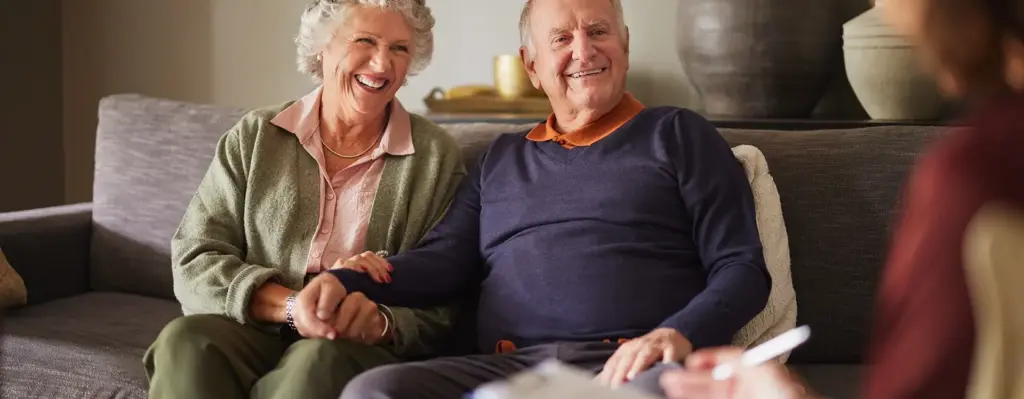 Das Bild zeigt ein älteres Pärchen, das auf einer Couch sitzt. Beide lächeln. Ihnen gegenüber sitzt eine weitere Person. Es ist eine Beratungssituation und wird im Bereich "Beratungseinsatz bei häuslicher Pflege" verwendet.