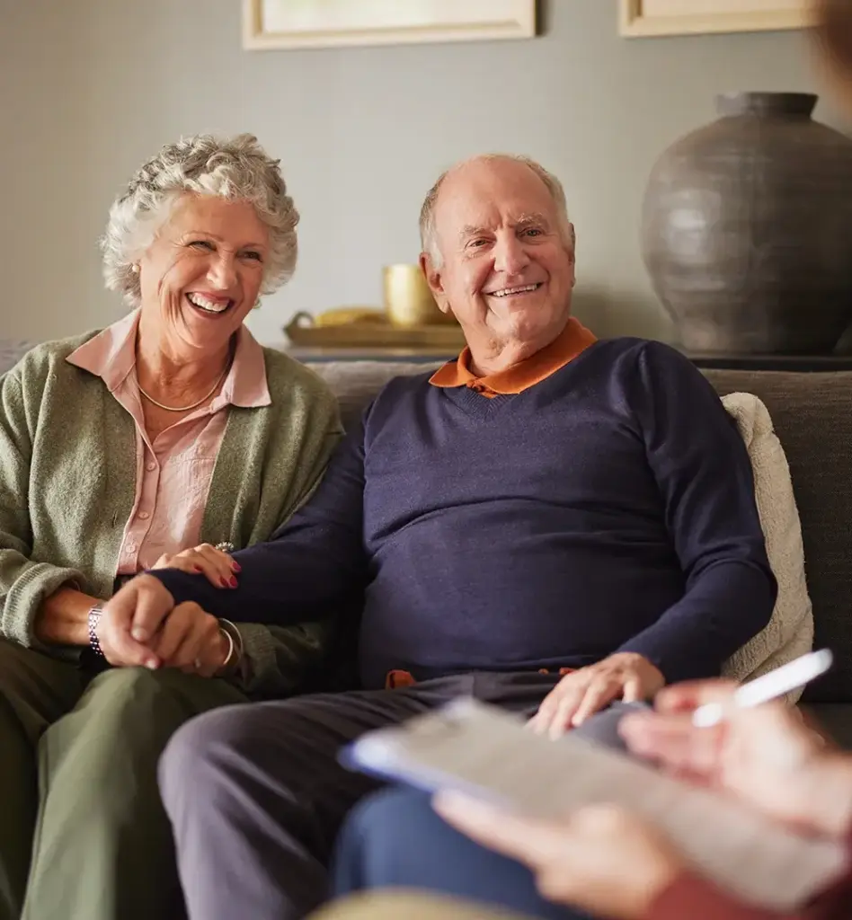 Das Bild zeigt ein älteres Pärchen, das auf einer Couch sitzt. Beide lächeln. Ihnen gegenüber sitzt eine weitere Person. Es ist eine Beratungssituation und wird im Bereich "Beratungseinsatz bei häuslicher Pflege" verwendet.