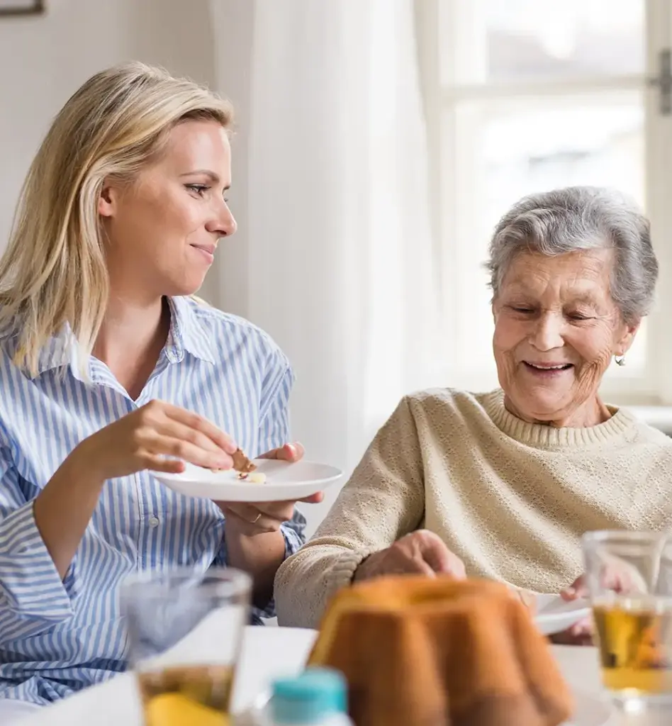 Das Bild zeigt eine ältere Frau, die lacht. Sie sitzt an einem Tisch und hält einen Teller mit der Hand fest. Neben ihr sitzt eine jüngere Frau, die ebenfalls einen Teller hält und ein Stück Kuchen ist. Das Bild wird im Bereich "Betreuungs- und Entlastungsleistungen" verwendet.