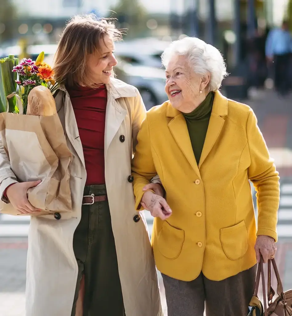 Das Bild zeigt eine ältere Frau, die von einer jüngeren Frau begleitet wird. Die jüngere Frau trägt zusätzlich Einkäufe in einer Papiertüte. Das Bild wird im Bereich "Kombination von Sach- und Geldleistungen" verwendet.