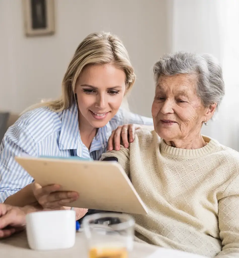 Das Bild zeigt eine ältere Frau im Rollstuhl, die ein Klemmbrett in der Handhält. Neben ihr kniet eine jüngere Frau. Zusammen blicken sie auf das Klemmbrett. Das Bild wird im Bereich "Kurzzeitpflege" verwendet.