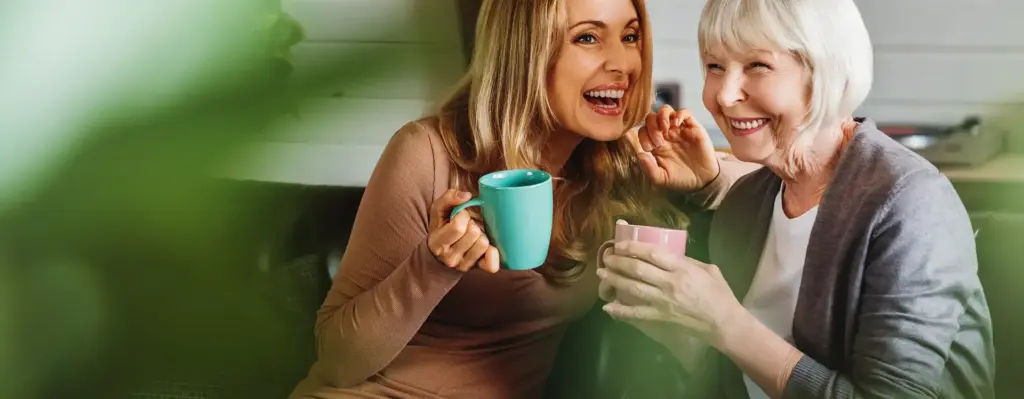 Das Bild zeigt eine jüngere Frau und eine ältere Frau, die zusammen auf einer Couch sitzen. Beide Frauen halten eine Tasse in der Hand und lachen. Sie scheinen sich zu unterhalten. Das Bild wird beim Thema "Patientenverfügung, Vorsorgevollmacht, Betreuungsrecht" verwendet.