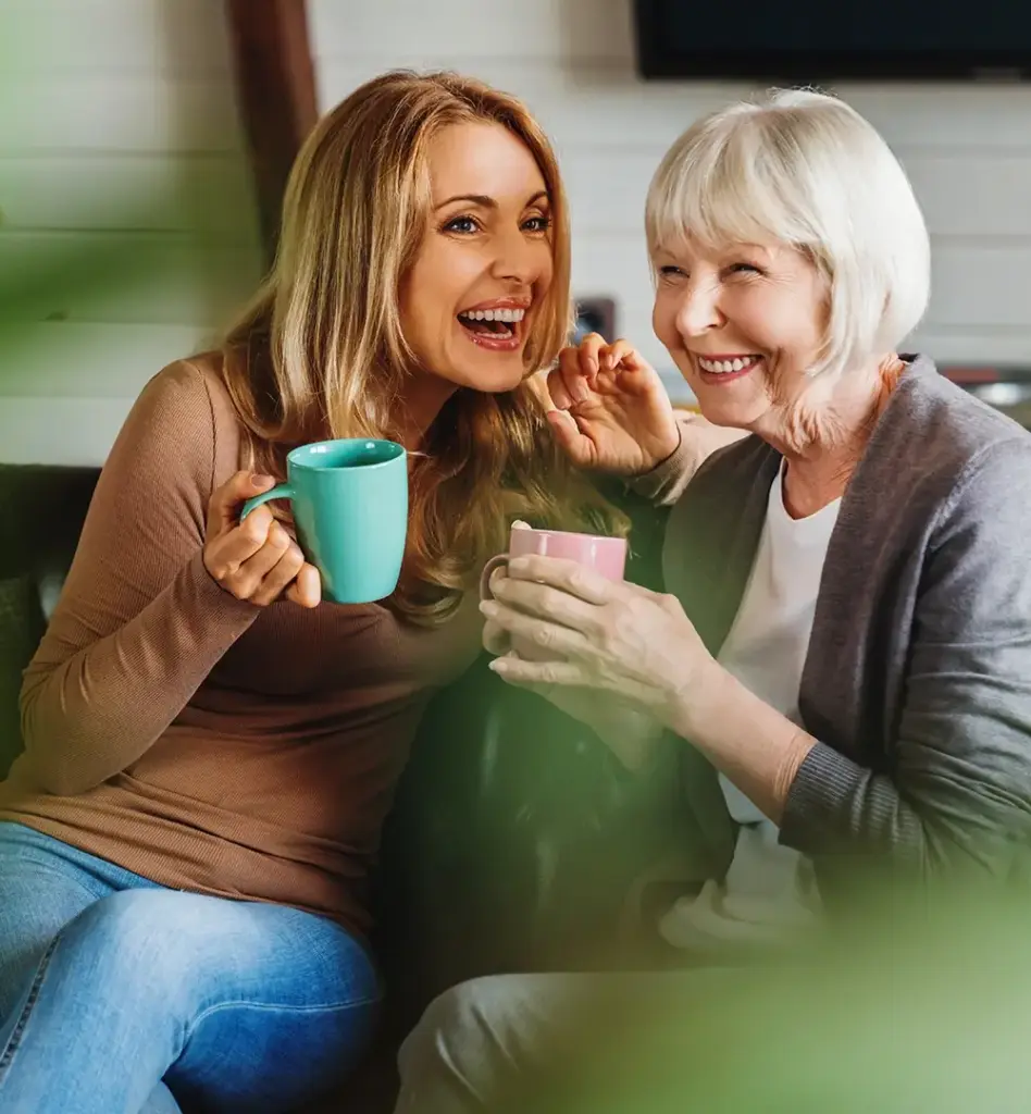 Das Bild zeigt eine jüngere Frau und eine ältere Frau, die zusammen auf einer Couch sitzen. Beide Frauen halten eine Tasse in der Hand und lachen. Sie scheinen sich zu unterhalten. Das Bild wird beim Thema "Patientenverfügung, Vorsorgevollmacht, Betreuungsrecht" verwendet.