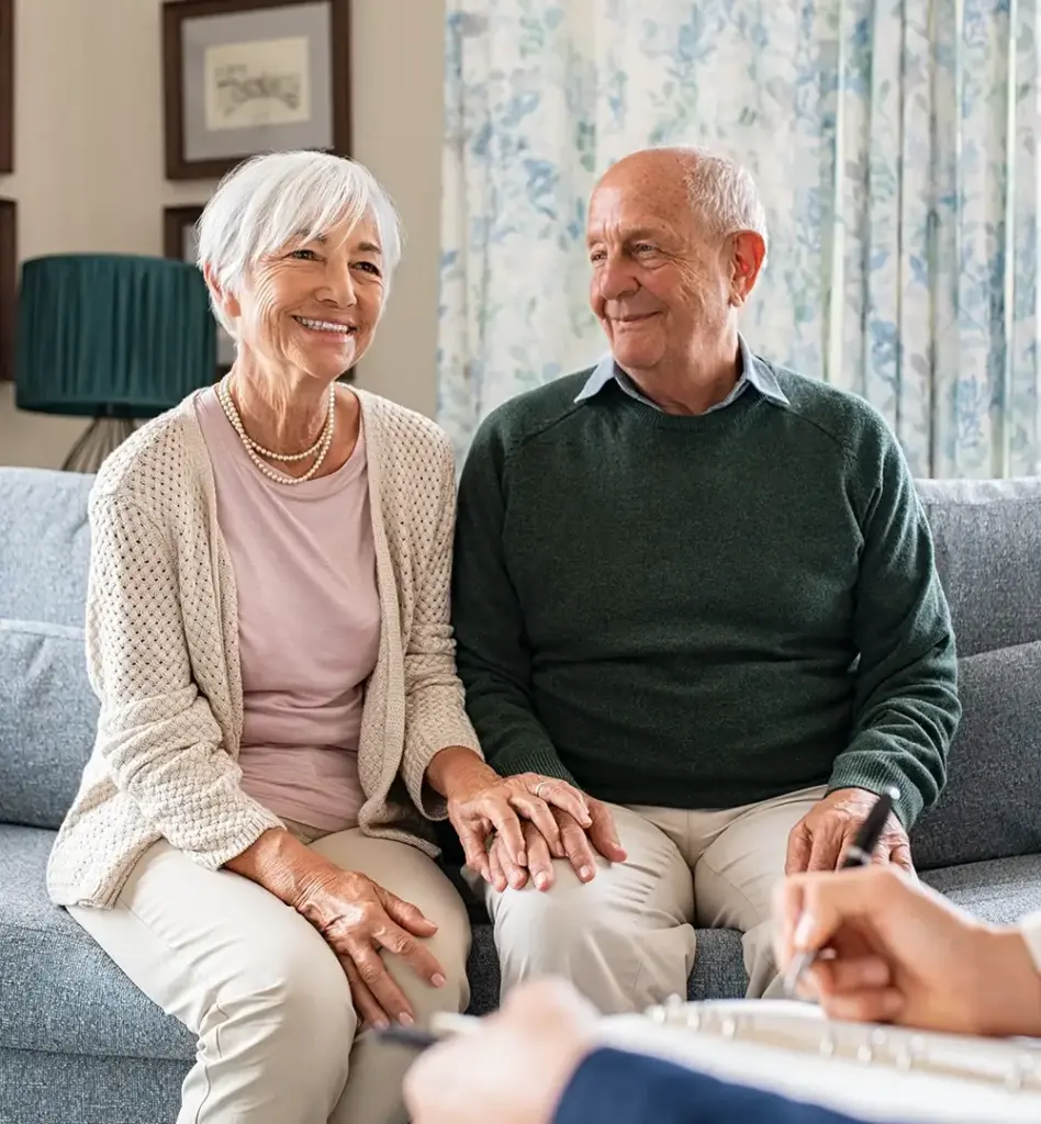 Das Bild zeigt ein älteres Pärchen, das auf einer Couch sitzt. Der Mann schaut seine Frau an. Ihnen gegenüber sitzt eine weitere Person. Es ist eine Beratungssituation und wird auf der Leistungsseite "Pflegeberatung und Pflegestützpunkte" genutzt.
