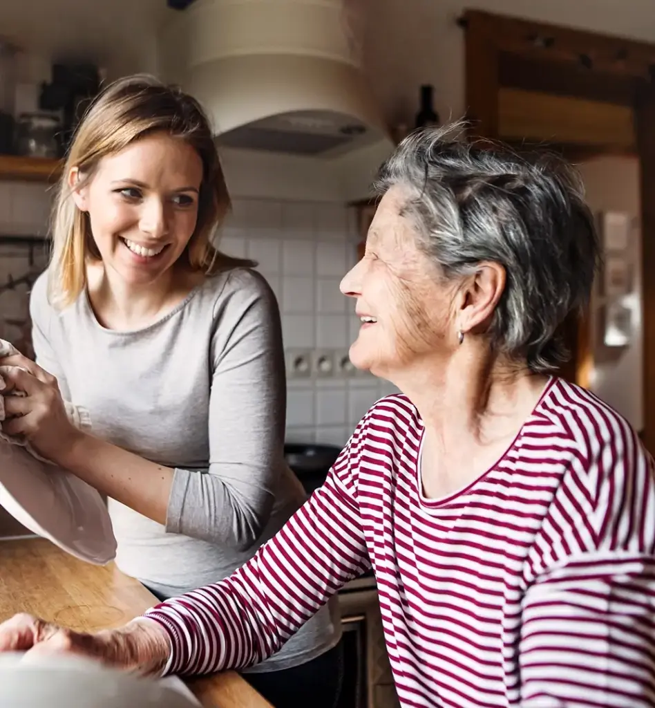 Das Bild zeigt eine ältere Frau in einem rot-weiß gestreiften Shirt. Sie steht in der Küche an einer Spüle. Neben ihr steht eine jüngere Frau, die einen Teller abtrocknet. Das Bild wird im Bereich "Pflegegeld für sogenannte Arbeitgeber­modelle" verwendet.