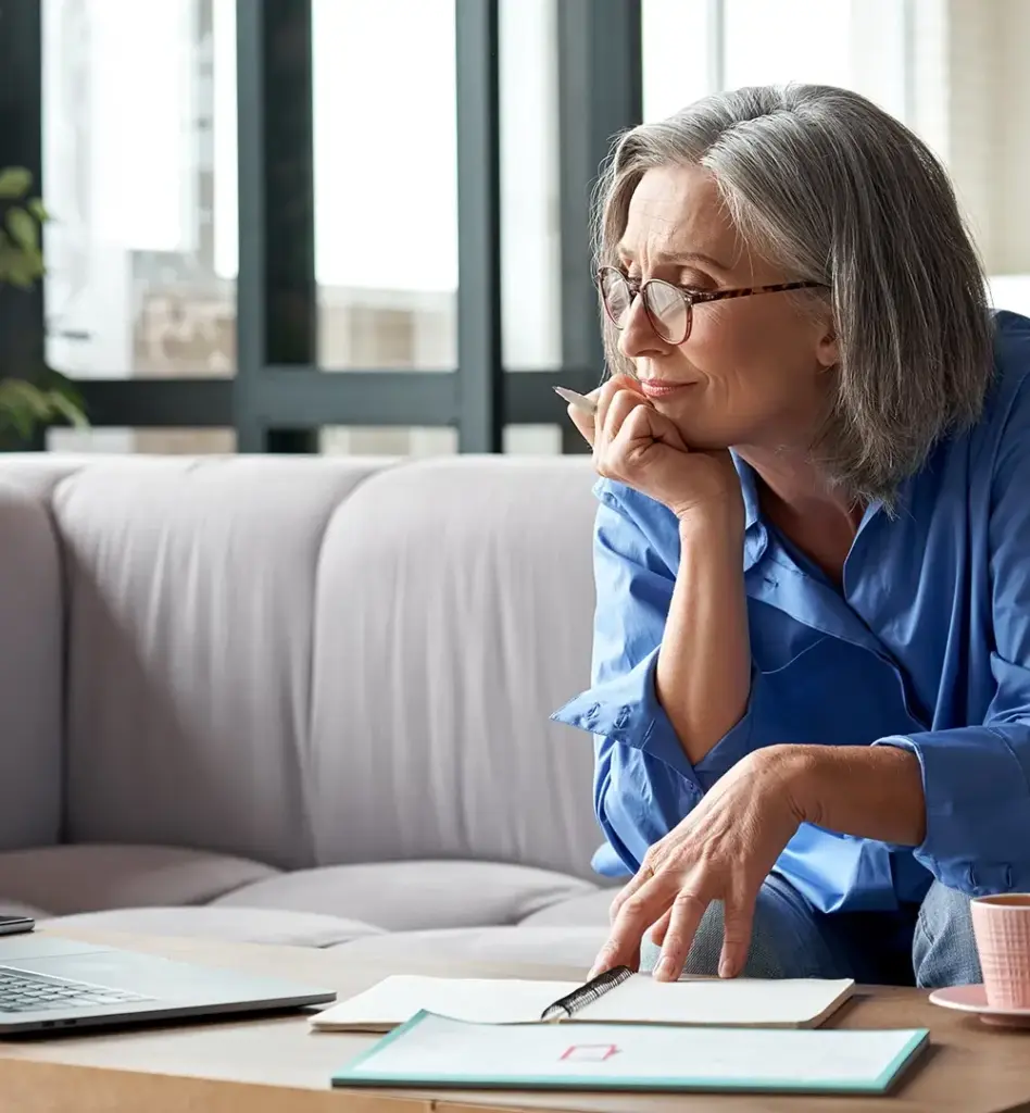 Das Bild zeigt eine Frau, die auf einer Couch sitzt. Sie lehnt nach vorne, stützt ihren rechten Ellenbogen auf ihr Knie und ihre Hand mit Stift ans Kinn. Sie schaut auf einen Laptop, der auf auf dem Wohnzimmertisch steht. Das Bild wird für die Leistung "Pflegekurse" genutzt.