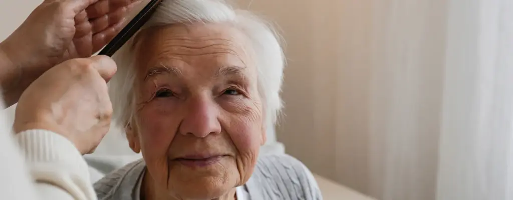Eine ältere Frau blickt schräg nach oben zu einer Person, die ihr mit einem Kamm die Haare kämmt. Die weitere Person ist nur mit ihren Händchen zu sehen. Das Bild wird im Bereich "Pflegeunterstützungsgeld" verwendet.