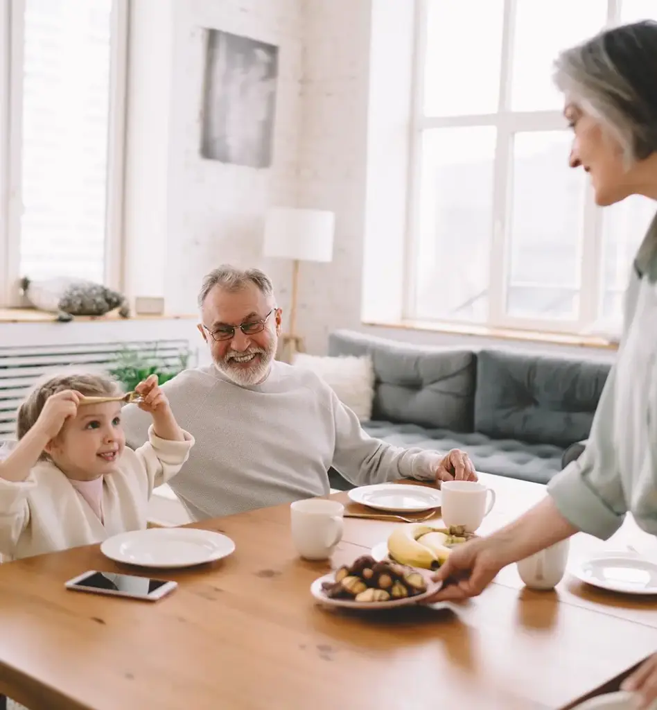 Das Bild zeigt einen älteren Mann, der am Esstisch sitzt. Neben ihm sitzt ein kleines Mädchen - die Enkelin. Eine ältere Frau stellt Kekse auf den Tisch. Das Bild wird im Bereich "Pflegezeit/Familienpflegezeit" verwendet.