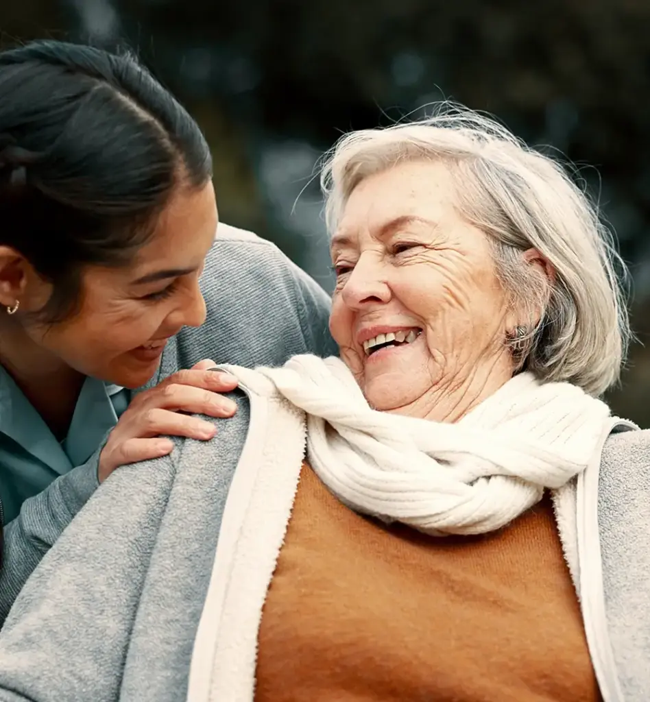Das Bild zeigt eine ältere Frau im Rollstuhl, wobei sie nur von Kopf bis zur Brust zu sehen ist. Von hinten lehnt eine jüngere Frau über ihre Schulter und lächelt sie an. Das Bild wird im Bereich "Pflegevorsorgefonds – Private Pflegevorsorge" verwendet.