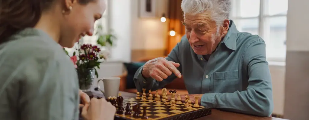 Das Bild zeigt einen älteren Mann, der am Tisch sitzt, auf dem ein Schachbrett liegt. Ihm gegenüber sitzt eine jüngere Frau. Man sieht außerdem eine Tasse auf dem Tisch und Blumen. Das Bild wird im Bereich "Sachleistung bei häuslicher Pflege" verwendet.