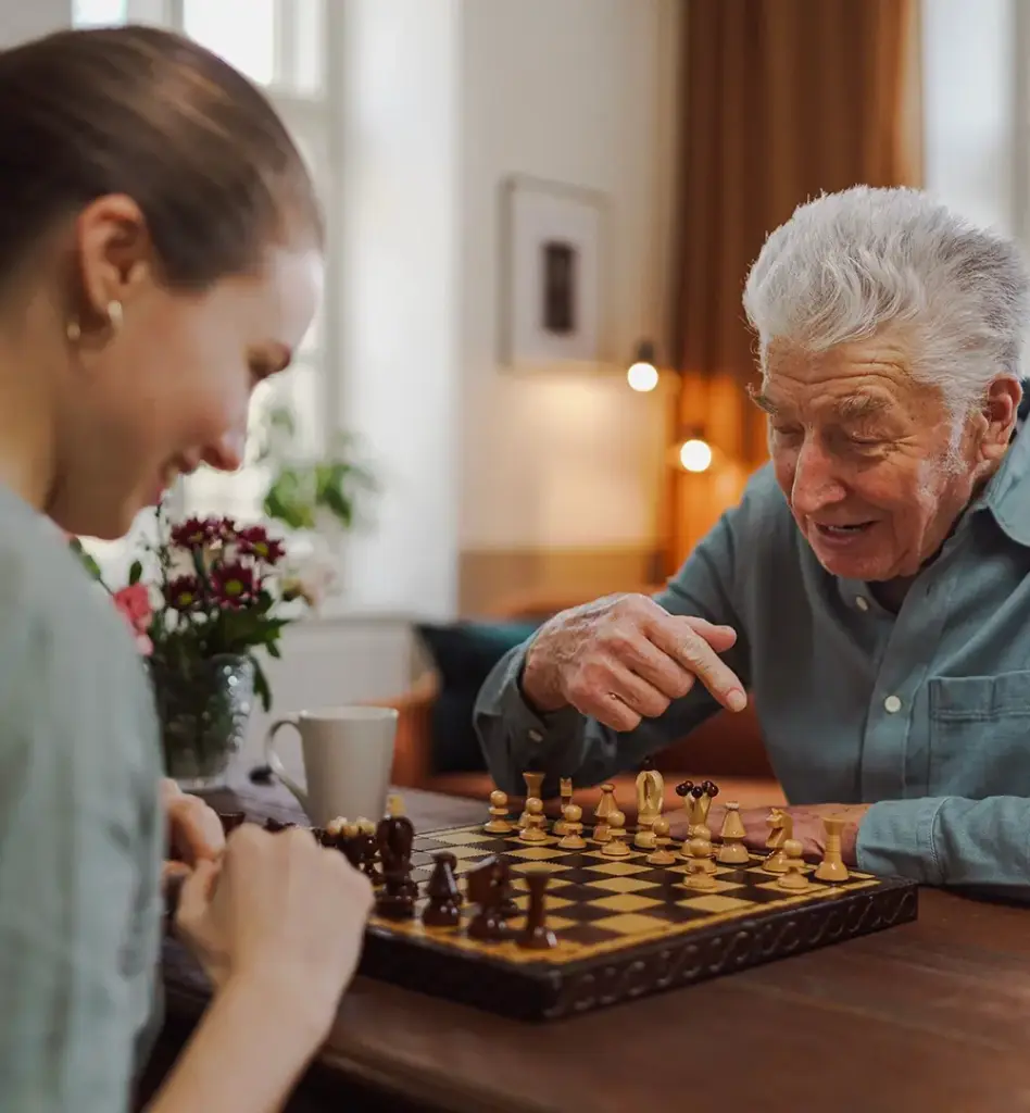 Das Bild zeigt einen älteren Mann, der am Tisch sitzt, auf dem ein Schachbrett liegt. Ihm gegenüber sitzt eine jüngere Frau. Man sieht außerdem eine Tasse auf dem Tisch und Blumen. Das Bild wird im Bereich "Sachleistung bei häuslicher Pflege" verwendet.