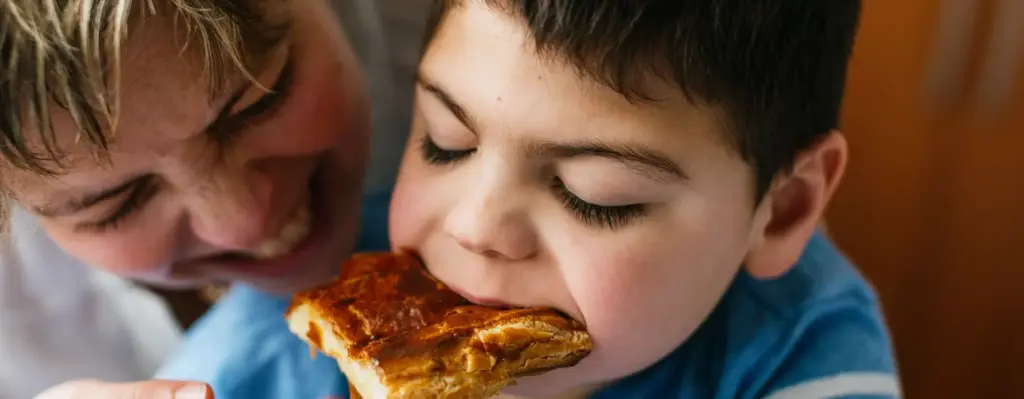 Das Bild zeigt einen Jungen mit Mikrodeletionssyndrom, der mit geschlossenen Augen, genüsslich in ein Stück Pizza beißt. Er wird von seiner lachenden Mutter umarmt. Das Bild wird im Bereich "Soziale Sicherung der Pflegepersonen" verwendet.