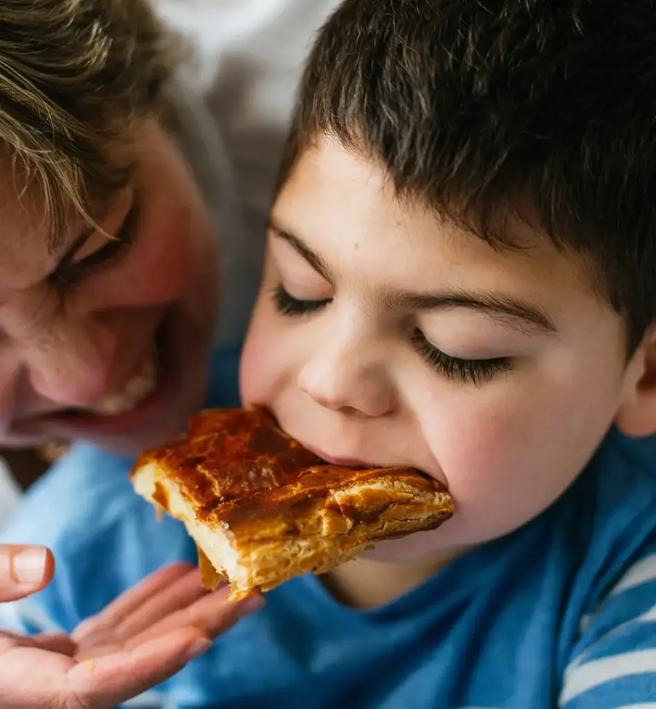 Das Bild zeigt einen Jungen mit Mikrodeletionssyndrom, der mit geschlossenen Augen, genüsslich in ein Stück Pizza beißt. Er wird von seiner lachenden Mutter umarmt. Das Bild wird im Bereich "Soziale Sicherung der Pflegepersonen" verwendet.