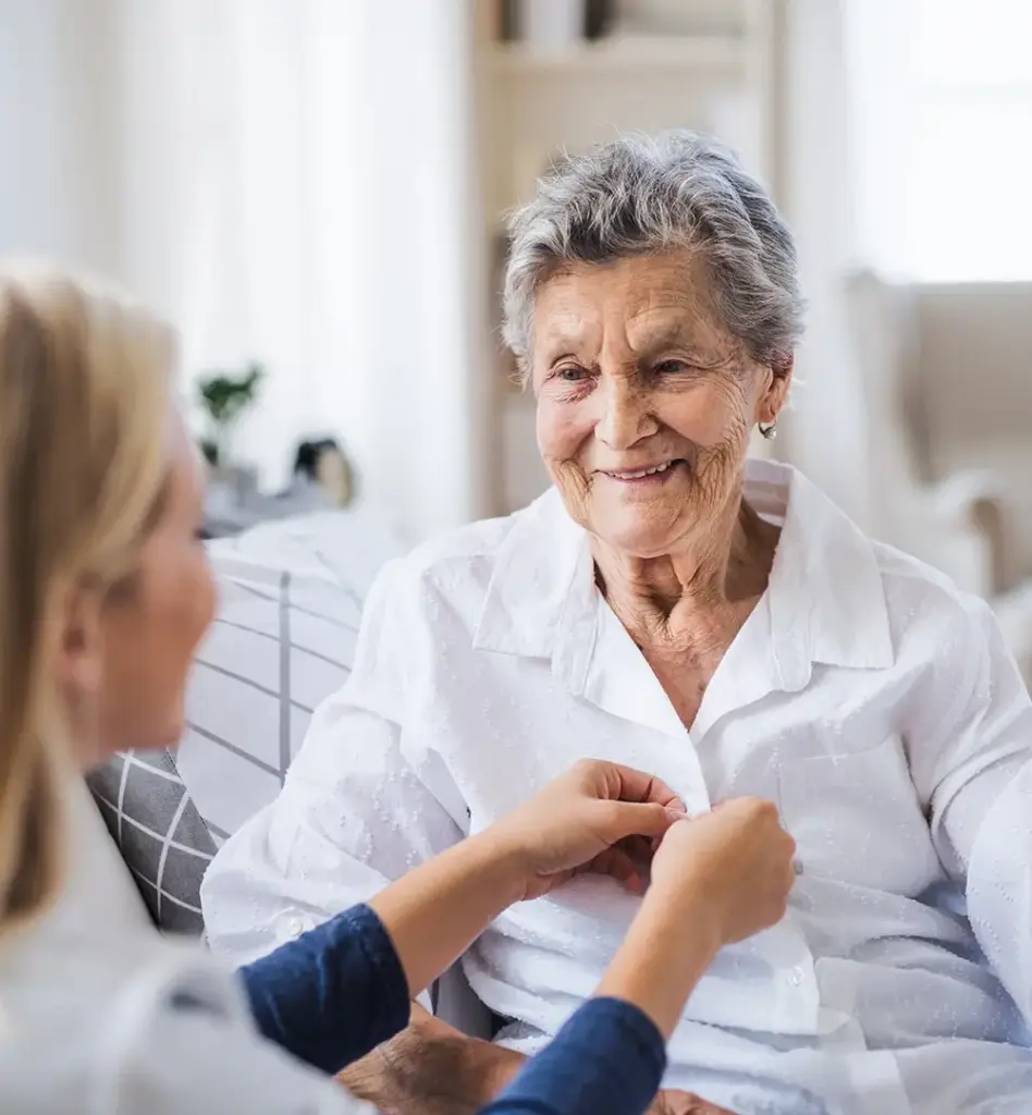 Das Bild zeigt eine ältere Frau, die auf einem Bett sitzt. Eine jüngere Frau knöpft ihr den Pyjama zu. Das Bild wird im Bereich "Teilstationäre Tages- und Nachtpflege" verwendet.