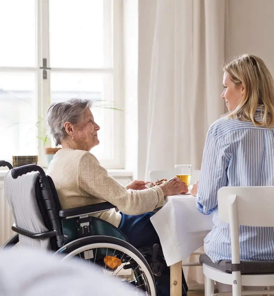 Das Bild zeigt eine ältere Frau, die lächelt. Sie sitzt in einem Rollstuhl an einem Tisch, auf dem Gläser und Teller stehen. Neben ihr sitzt eine jüngere Frau mit Rücken zur Kamera. Beide Personen schauen sich an. Das Bild wird im Bereich "Verhinderungspflege" verwendet.