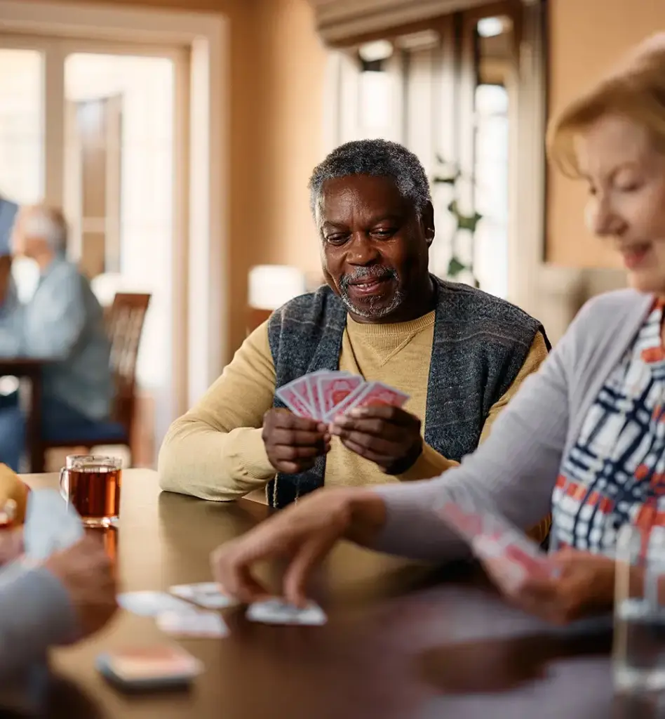 Das Bild zeigt eine Gruppe von älteren Menschen, die zusammen Kartenspielen. Im Fokus des Bildes ist ein älterer Mann, der sechs Spielkarten auf der Hand hält. Das Bild wird im Bereich "Wohngruppen (ambulant betreute)" verwendet.