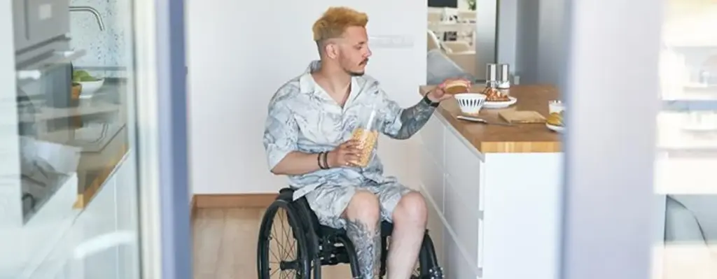 Ein Mann sitzt im Rollstuhl. An einer Küchenzeile bereitet er sich etwas zu essen vor. In einer Hand hält er ein Glas. Das Bild wird im Bereich "Wohnungsanpassung" verwendet.