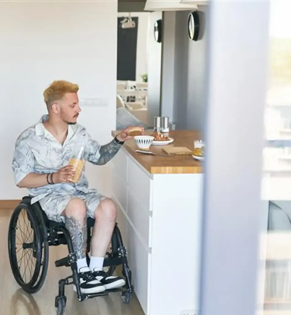 Ein Mann sitzt im Rollstuhl. An einer Küchenzeile bereitet er sich etwas zu essen vor. In einer Hand hält er ein Glas. Das Bild wird im Bereich "Wohnungsanpassung" verwendet.