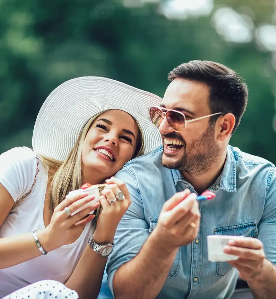 Eine Frau mit weißem Sonnenhut lehnt an die Schultern eines Mannes. Beide lachen und halten einen Becher mit Eis in der Hand.