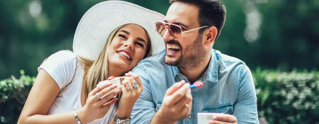 Eine Frau mit weißem Sonnenhut lehnt an die Schultern eines Mannes. Beide lachen und halten einen Becher mit Eis in der Hand.