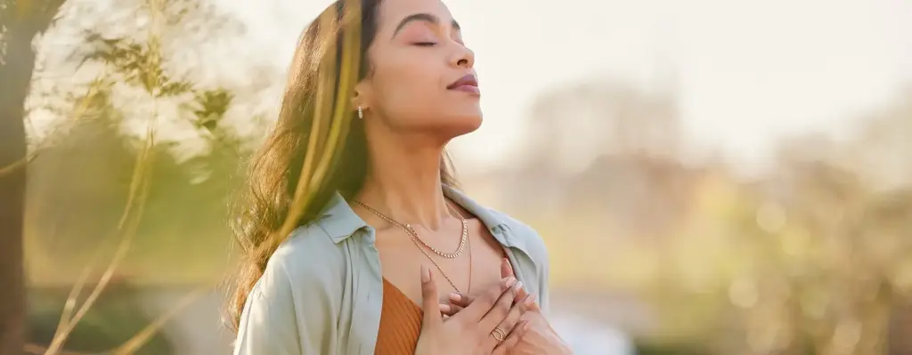 Eine Frau steht in der Natur, hält die Hände auf der Brust, hat die Augen geschlossen und scheint zu meditieren.