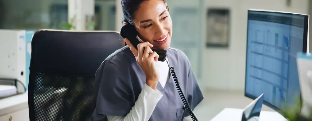 Eine im Krankenhaus arbeitende Frau sitzt am Schreibtisch vor einem Bildschirm und telefoniert mit einem Schnurtelefon. Das Bild wird im Bereich "24/7 Arzthotline" verwendet.