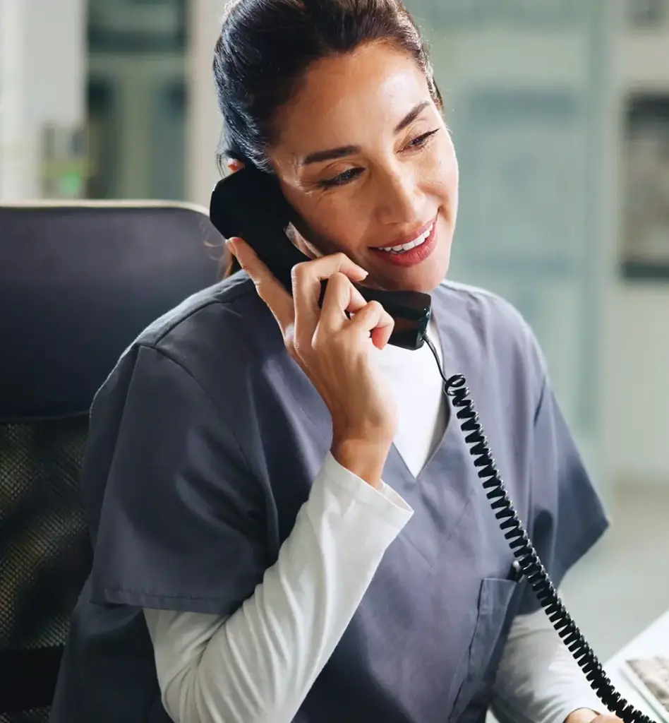 Eine im Krankenhaus arbeitende Frau sitzt am Schreibtisch vor einem Bildschirm und telefoniert mit einem Schnurtelefon. Das Bild wird im Bereich "24/7 Arzthotline" verwendet.