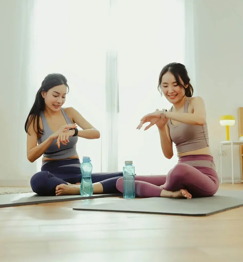 Zwei Frauen sitzen in einem Zimmer mit Bett auf Fitnessmatten. Sie schauen jeweils auf ihre Sportuhr. Vor ihnen steht jeweils eine Wasserflasche. Das Bild wird im Bereich "Aktiv und gesund" verwendet.