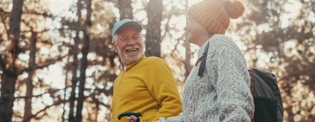 Ein älteres Paar blickt sich an und lacht. Sie sind mit Walkingstöcken bzw. Wanderstöcken unterwegs. Der Mann trägt einen gelben Pullover und eine Cap. Die Frau trägt einen grauen Strickpullover und eine orangefarbene Pudelmütze. Das Bild wird für die Zielgruppenseite Best Ager zum Thema "Aktiv vorsorgen – mit Zuschuss für Ihre Gesundheitskurse" genutzt.