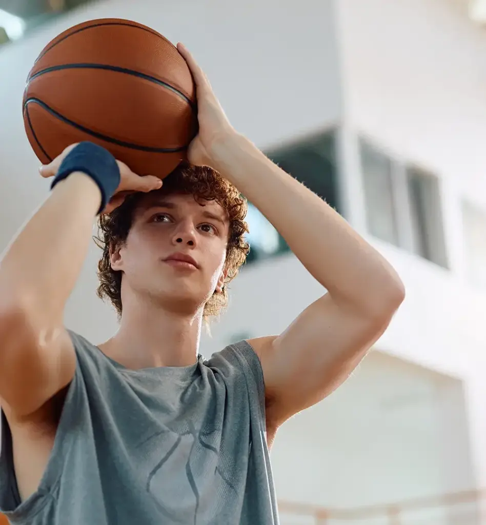 Ein junger Mann in Sportkleidung hält einen Basketball mit beiden Händen hoch und setzt zum Wurf an. Das Bild wird im Bereich "Aktivbonus Präventionskurs" verwendet.