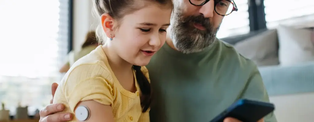 Ein Mädchen sitzt auf dem Schoß ihres Vaters. Sie hat einen Diabetes Zuckersensor am Oberarm und schaut zusammen mit ihrem Vater auf ein Smartphone. Das Bild wird im Bereich "Digitale Gesundheitsbegleiter für Ihren Familienalltag" verwendet.
