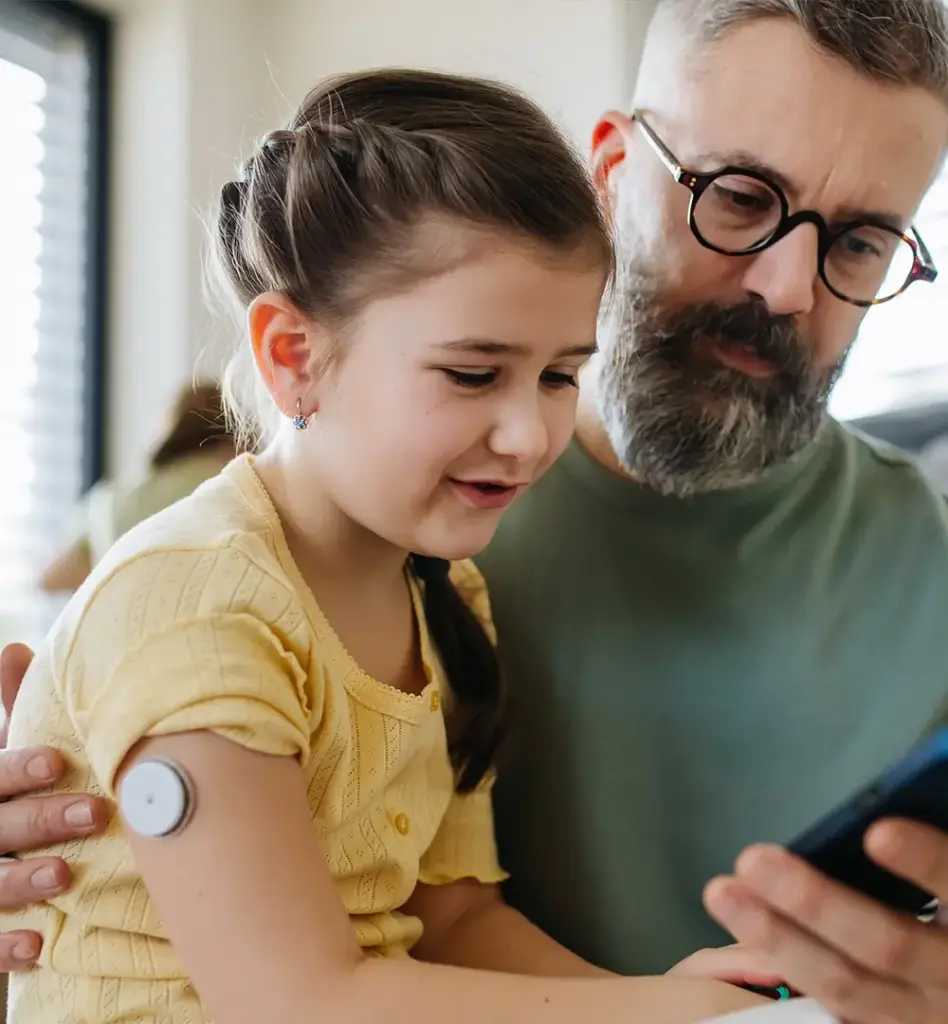 Ein Mädchen sitzt auf dem Schoß ihres Vaters. Sie hat einen Diabetes Zuckersensor am Oberarm und schaut zusammen mit ihrem Vater auf ein Smartphone. Das Bild wird im Bereich "Digitale Gesundheitsbegleiter für Ihren Familienalltag" verwendet.