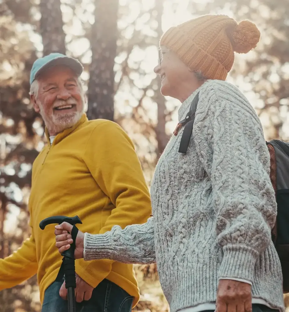 Ein älteres Paar blickt sich an und lacht. Sie sind mit Walkingstöcken bzw. Wanderstöcken unterwegs. Der Mann trägt einen gelben Pullover und eine Cap. Die Frau trägt einen grauen Strickpullover und eine orangefarbene Pudelmütze. Das Bild wird für die Zielgruppenseite Best Ager zum Thema "Aktiv vorsorgen – mit Zuschuss für Ihre Gesundheitskurse" genutzt.