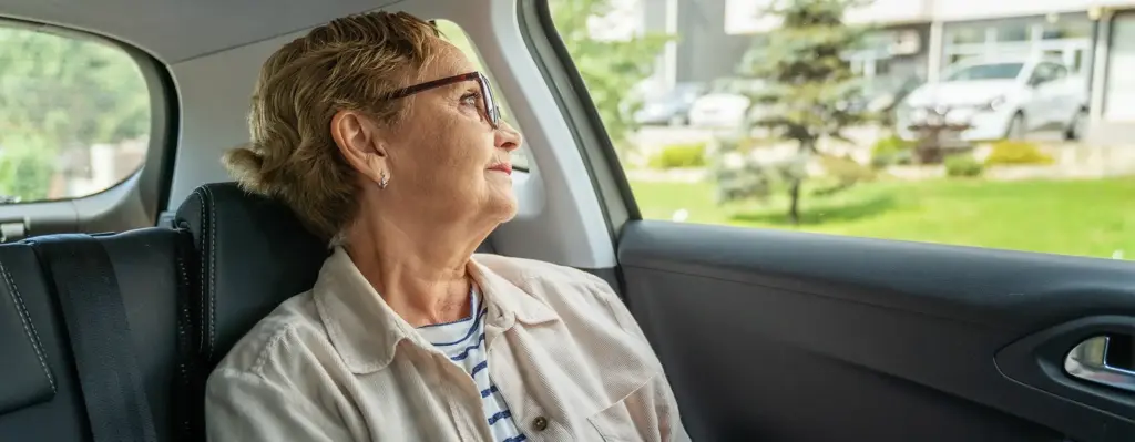 Eine Frau sitzt in einem Auto auf der Rückband hinter dem Fahrer und blickt aus dem Fenster. Das Bild wird im Bereich "Fahr- und Transportkosten" verwendet.
