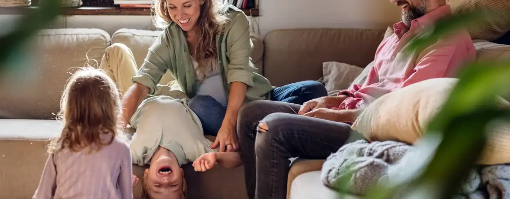 Eine Frau sitzt auf der Couch. Sie hält einen Jungen fest, der kopfüber von der Couch hängt. Am Bildrand sitzt ein Mann. Das Bild wird im Bereich "Fit von klein auf - Kindergartenkonzept" verwendet.