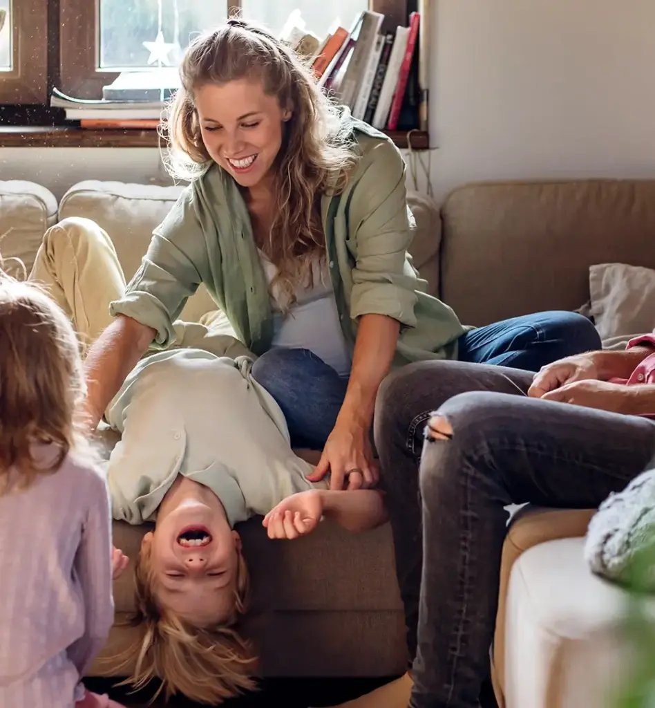 Eine Frau sitzt auf der Couch. Sie hält einen Jungen fest, der kopfüber von der Couch hängt. Am Bildrand sitzt ein Mann. Das Bild wird im Bereich "Fit von klein auf - Kindergartenkonzept" verwendet.