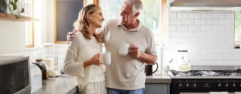 Das Bild zeigt einen älteren Mann und eine Frau in einer Küche. Der Mann hat seinen Arm um die Schultern der Frau gelegt. Beide halten Kaffeetassen in der Hand und lächeln sich an. Das Bild wird im Bereich "Gesund bleiben und Bonus sichern" verwendet.